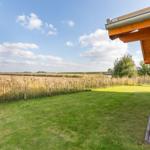 Hinter dem Holzhaus erstreckt sich ein grüner Rasenbereich mit einem Zaun und Blick auf Felder.