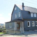 Haus mit dunklem Schieferdach und weißen Fenstern an einer Straße mit Gartenzaun.