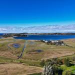 Aerielansicht eines Sees mit Uferbereichen und Grünflächen unter blauem Himmel.