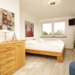 Schlafzimmer mit Holzbett, Schrank und Fenster. Wandbilder und Pflanze sichtbar.