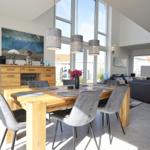 Essbereich mit Holztisch und grauen Stühlen. Große Fenster und Wohnbereich im Hintergrund.