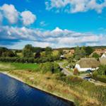 Haus am Fluss mit grünen Wiesen und Wald im Hintergrund unter blauem Himmel.