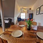 Holztisch mit Stühlen, Blumen und Geschirr. Im Hintergrund Küche und Wohnbereich.