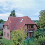 Zweistöckiges Haus mit rotem Dach und Balkon in grüner Umgebung.