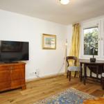 Zimmer mit Holzboden, TV auf Kommode, Tisch mit zwei Stühlen und Fenster mit Vorhängen.