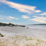 Strand mit Sand, Boot und Holzsteg am Wasser unter blauem Himmel.