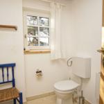 Badezimmer mit Toilette, blauer Stuhl und Fenster mit Vorhang.