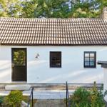 Wohnhaus mit weißer Fassade, schwarzem Eingang und zwei Fenstern, umgeben von Grün.