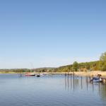 Stille See mit Booten und Holzstegen. Grün bewachsener Uferbereich mit Häusern im Hintergrund.