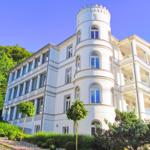 Weißes Gebäude mit Turm und Balkonen, umgeben von Grünflächen und Bäumen.