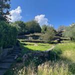 Path ascends through green meadows and stone walls under blue sky.