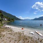 Gravel beach, lake, and mountains. People swimming and sunbathing.