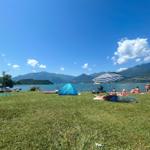 Green shore with people, tent, and beach chairs by the lake.