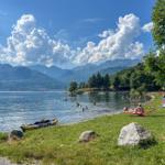 Lake shore with swimmers, boat, and mountains in the background.