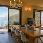 Esszimmer mit Blick auf See und Berge. Holztisch, Stühle, TV und Balkon.