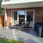 Terrasse mit Tisch, Stühlen und Grill. Glasfront führt ins Haus. Holzumrandung und Grasfläche.