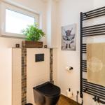 Badezimmer mit Toilette, Heizkörper und Fenster. Wandbild und Pflanze sichtbar.