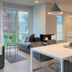 Moderne Wohnküche mit Esstisch, Sofa und Kaminofen. Große Fenster mit Blick auf Garten.