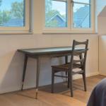 Schreibtisch und Stuhl vor Fenster mit Blick auf Dächer und Bäume.