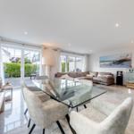 Ein Essbereich mit Glas-Tisch und Stühlen. Im Hintergrund ein Wohnbereich mit Sofa und großen Fenstern.