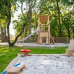 Playground with sandbox, slide, and wooden play equipment in greenery.