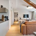 Apartment with kitchen, living area, and wooden beam. Sofa, TV, and window visible.