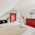 Schlafzimmer mit rotem Schrank und Kommode, Bett mit kariertem Bettzeug, Blick in die Küche.