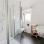 Badezimmer mit Dusche, Toilette und Waschbecken. Fenster mit Rollladen und Holzregal.