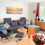 Wohnzimmer mit grauem Sofa, Ledersessel und Holztisch. Fenster mit roten Vorhängen.