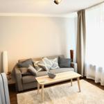 Ein Wohnzimmer mit grauem Sofa, Holztisch und Fenster mit Vorhängen.