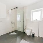 Badezimmer mit Dusche, Toilette und Fenster. Fliesenboden und weiße Wände.