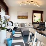 Living and dining area with sofa, chairs, and table. Windows and wall art visible.