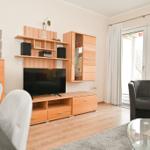 Wohnzimmer mit Holzmöbeln, Fernseher und Sitzgelegenheiten. Durchgang zur Terrasse mit Sicht auf die Umgebung.