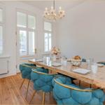 Esszimmer mit Holztisch, blauen Stühlen und Kronleuchter. Große Fenster und Holzboden.