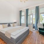 Ein Schlafzimmer mit Doppelbett, grünem Sessel und Holzboden. Fenster mit blauen Vorhängen.