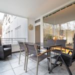 Terrasse mit Tisch, Stühlen und Sessel. Glaswände und Blick auf Gebäude.