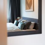 Ein Bett mit Kissen und Decke ist im Spiegel zu sehen. Dunkle Vorhänge und Wandbild sind sichtbar.