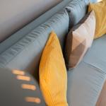 Ein hellblauer Sofa mit gelben und grauen Kissen. Holzboden und Fensterblick.