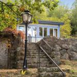 Ein blaues Haus mit Fenstern, Zugang über Steintreppe und Laterne. Umgeben von Bäumen und Steinmauer.