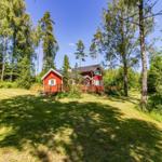 Rotes Holzhaus mit Terrasse im Wald. Grünflächen und hohe Bäume umgeben das Haus.