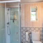 Badezimmer mit Dusche, Toilette und Fenster. Wände aus Holz und Mosaikfliesen.