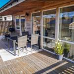Terrasse mit Sitzgruppe, Grill und Pflanze vor Holzhaus mit Glasfront.