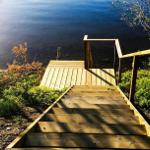 Holzstufen führen zu einer Terrasse am See. Umgeben von Bäumen und Grün.