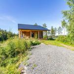 Wooden cabin with porch, surrounded by forest and grassland under blue sky.