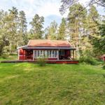 Red wooden house with terrace in the forest, green lawn and tall trees surround.