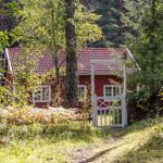Red wooden house with white gate in the forest. Windows and roof are visible.