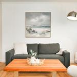 Wohnzimmer mit Sofa, Couchtisch und Sessel. Wandbild zeigt Strand. Tür führt ins Schlafzimmer.