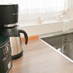 Küchenzeile mit Kaffeemaschine, Kaffeekanne und Herd. Fenster mit weißen Vorhängen.