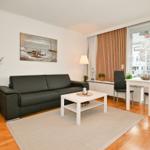 Wohn- und Essbereich mit Sofa, Tisch und Stühlen. Holzboden und Fenster mit Jalousien.