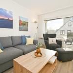 Wohnzimmer mit Sofa, Sessel und Tisch. Blick auf Balkon mit Stuhl. Wandbilder und Holzboden.